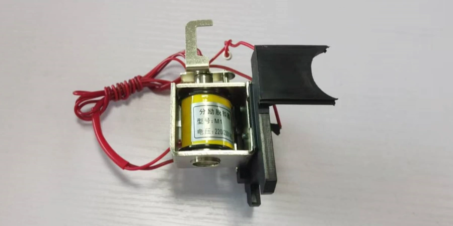 A close-up view of a shunt trip component for a Molded Case Circuit Breaker (MCCB), featuring a yellow and black electromagnetic coil with a label indicating "220V 0.1A," mounted on a metal frame. A red wire with a coiled section extends from the device, and a black plastic cover is partially attached, all placed on a light-colored surface.