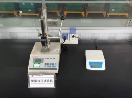 A laboratory workbench displaying, on the left, a Spring Tension and Compression Testing Machine with a digital display, control buttons, and manual lever for force application, and on the right, a compact digital scale with an LCD readout. A bilingual label (Chinese and English) is positioned in front of the machine, with industrial shelving and green storage bins visible in the background.