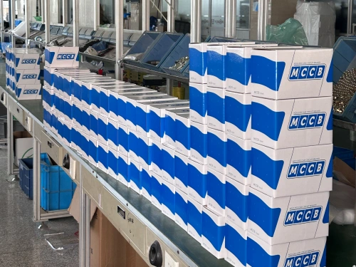 A photo showing a long row of white cardboard boxes with blue "MCCB" labels stacked in multiple layers along an assembly line workstation. The boxes vary in height arrangement, with blue bins, shelving units, and factory equipment visible in the background, indicating an active production environment.