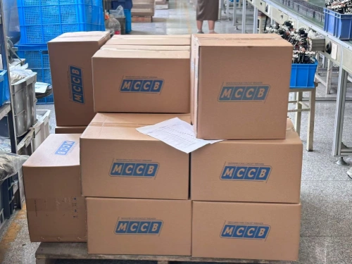 A photo showing a stack of cardboard boxes labeled "MCCB" neatly arranged on a pallet, stacked in multiple layers with a piece of paper placed on top of the middle stack. The boxes are positioned beside an assembly line, with a conveyor belt holding blue bins and a person standing nearby visible in the background, along with industrial shelving and equipment.