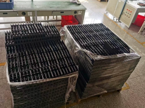 A warehouse scene featuring two large pallets stacked with black MCCB (Molded Case Circuit Breaker) bases, wrapped in protective plastic. The pallets are placed on a tiled floor near a green worktable. Under the table, red plastic crates used for holding products is visible, with various equipment in the background.
