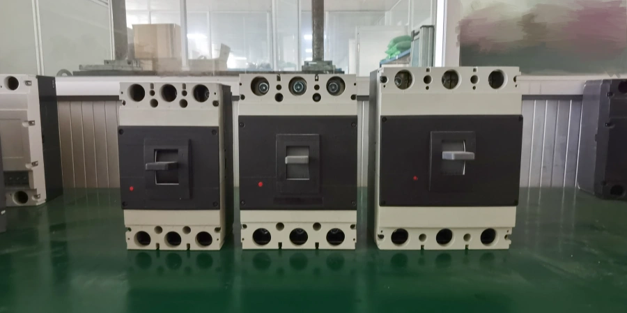 Three white and black molded case circuit breakers lined up on a green floor. From left to right: small, medium, and large size. Each has a gray handle, red indicator light, and multiple terminal holes on top and bottom.