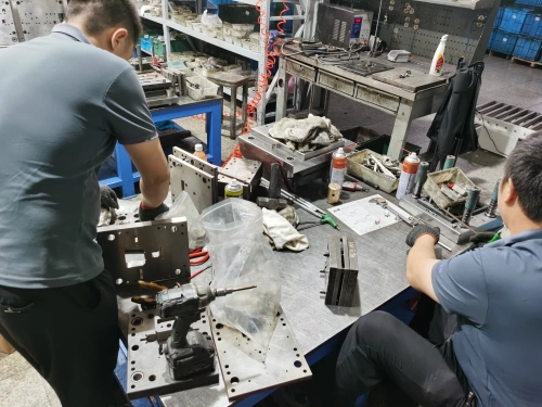 Two workers wearing blue shirts and gloves collaborate at a blue workbench in an industrial workshop, maintaining metal molds with intricate cutouts. A small drill is placed nearby. The table is cluttered with tools, a plastic bag, cans, and a green spray bottle. In the background, shelves with equipment, red coiled cords, a pegboard, and storage bins are visible.
