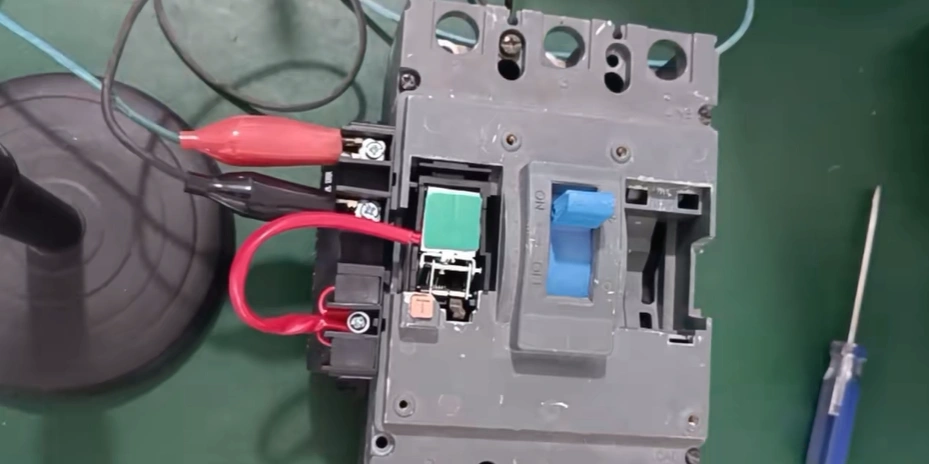 Close-up overhead view of a disassembled gray molded case circuit breaker (MCCB) on a green workbench during an undervoltage trip release trip test. The blue manual handle is flipped to the 