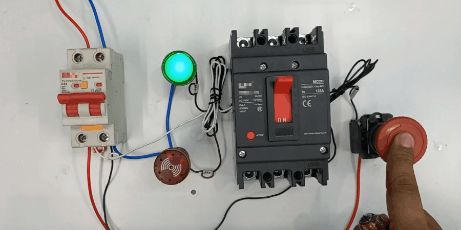 A demonstration setup on a white surface showing an emergency power-off system, featuring a red breaker connected via red and blue wires to a green illuminated indicator light and a red siren. A central black MCCB with a red switch is wired to a red emergency push button, which a finger is pressing to activate the shunt trip, illustrating remote circuit interruption for safety.