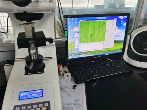 A laboratory workstation featuring a white Vickers Hardness Tester machine with a mounted metal sample under the diamond indenter, digital display showing zeroed measurements, and control panel. Adjacent is a Dell monitor running analysis software with a green fibrous material image and data graphs, a black keyboard, scattered blueprints and tools on the desk, and a white electric fan nearby, all in a brightly lit industrial room with window views.