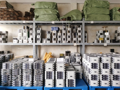 A warehouse shelf filled with neatly organized molded case circuit breakers (MCCBs) stacked in rows, with additional green bags and boxes stored on the upper shelves.