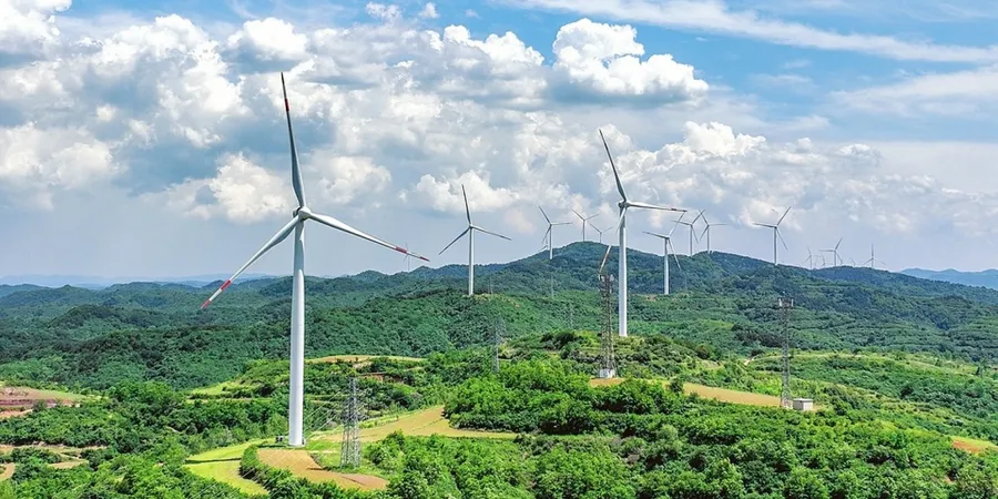 A scenic view of several large wind turbines standing on lush green hills under a partly cloudy blue sky, with more turbines visible in the distance on rolling mountains.