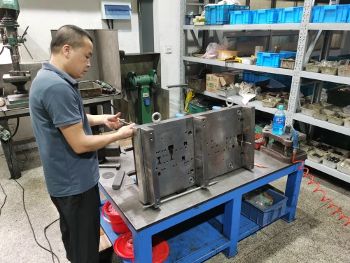 A worker in a blue shirt inspects large metal mold parts on a blue workbench in an industrial workshop. The parts have intricate cutouts and are placed next to tools and a red container. In the background, there are shelves with blue bins, a green drill press, and various equipment, with a coiled red power cord visible.