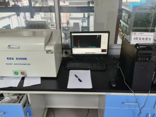 A laboratory desk featuring an EDX 8300B X-ray Fluorescence Spectrometer on the left, with a digital display and control button, positioned next to a computer monitor displaying a spectral graph, a keyboard, and a mouse. Papers and a pen are scattered on the desk, with a black computer tower and a labeled sign in Chinese on the right. Blue storage drawers are visible below the desk, and industrial equipment can be seen through the window in the background.