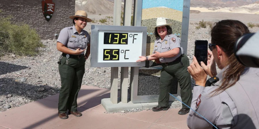 Two smiling female park rangers in tan uniforms and wide-brim hats stand next to a digital display showing 132°F (55°C) in a sunny desert setting with rocky terrain and a distant mountain. One ranger holds up the sign, while another takes a photo with her phone. This captures Phoenix, Arizona's extreme heat wave that broke a record with 19 consecutive days above 110°F in July 2023.