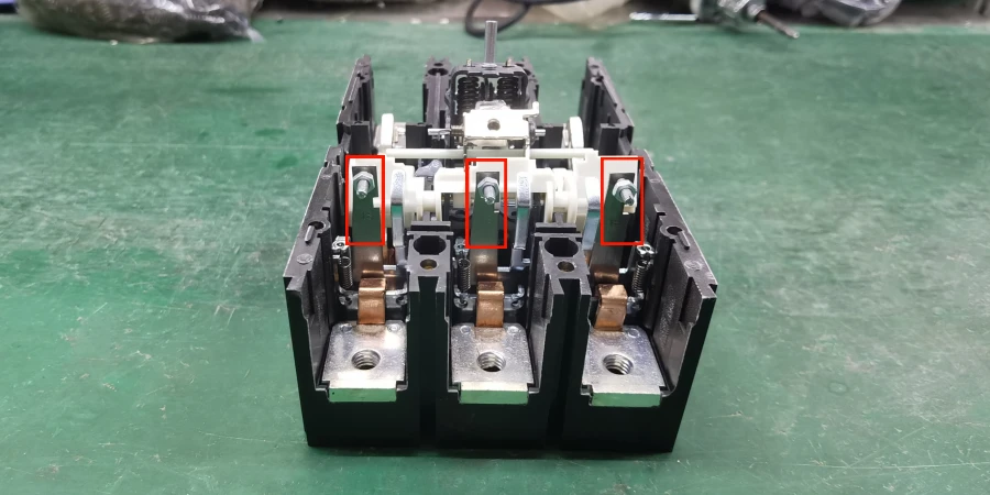 A molded case circuit breaker on a green workbench, showing three copper terminals and internal springs. The three bimetal strips are circled in red at the top.