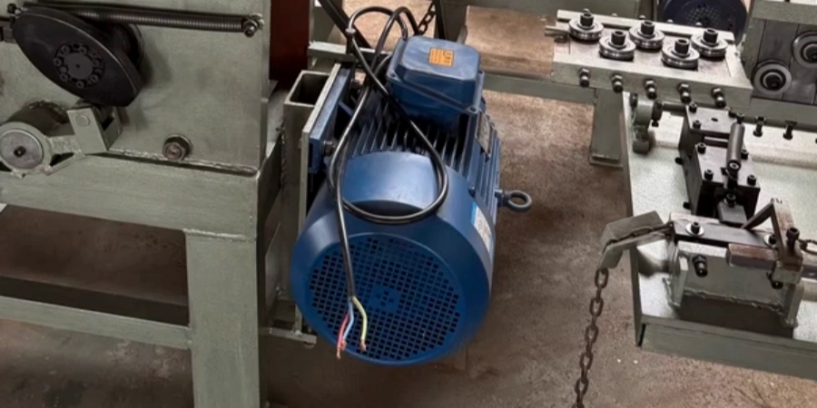 A blue industrial electric motor mounted on machinery, with its power cable entering the terminal box but several wires inside left exposed and unconnected. The motor is part of a mechanical setup with belts, pulleys, and chains visible around it.