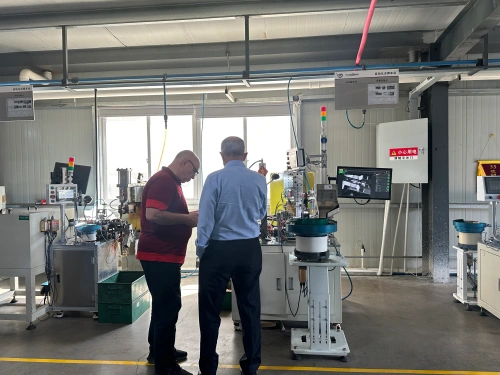 Two customers standing in a clean, well-lit factory, watching an automated riveting station where precision machines with monitors and feeding bowls rivet internal components of molded case circuit breakers (MCCBs) during a guided factory tour.