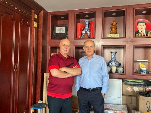 Two customers standing side by side in an office, one in a red polo shirt with arms crossed, the other in a light blue shirt with hands in pockets. Behind them is a dark wooden cabinet displaying blue-and-white porcelain vases, carved figurines, and framed artwork.