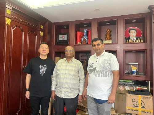 Three customers standing together in a wood-paneled office in front of a dark wooden display cabinet featuring blue-and-white porcelain vases, carved statues, and framed artwork, with some packed boxes visible on the right during a factory visit.