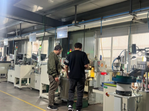Two customers touring the automated riveting workshop, observing high-precision riveting machines with green indicator lights actively joining internal components of molded case circuit breakers (MCCBs) on a modern factory assembly line.