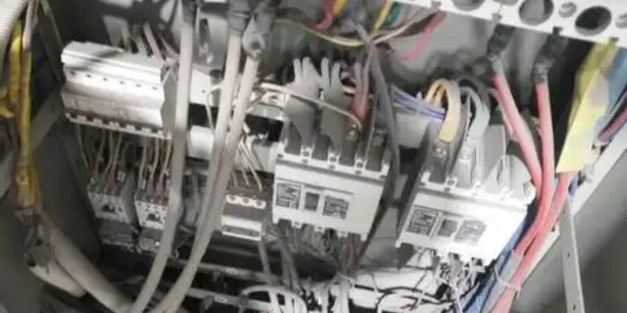 Cluttered and dusty electrical control cabinet filled with multiple circuit breakers, contactors, terminal blocks, and a chaotic bundle of colorful wires (red, yellow, blue, white) loosely tied and hanging, showing signs of poor maintenance and heavy dust buildup.