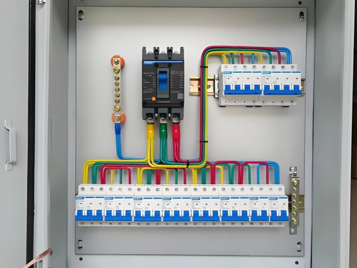 An open electrical panel showing a main mccb at the top, connected to colorful wires in red, yellow, green, and blue. Below are two rows of white mini circuit breakers lined up neatly, with wires running to a grounding bar on the left.