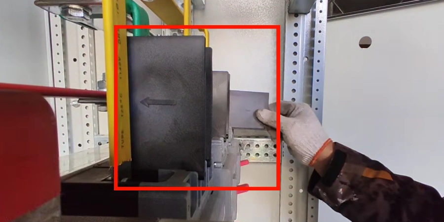 A gloved hand inserts a thin gray phase barrier into a black molded case circuit breaker mounted in a white electrical panel, with yellow and green busbars above.