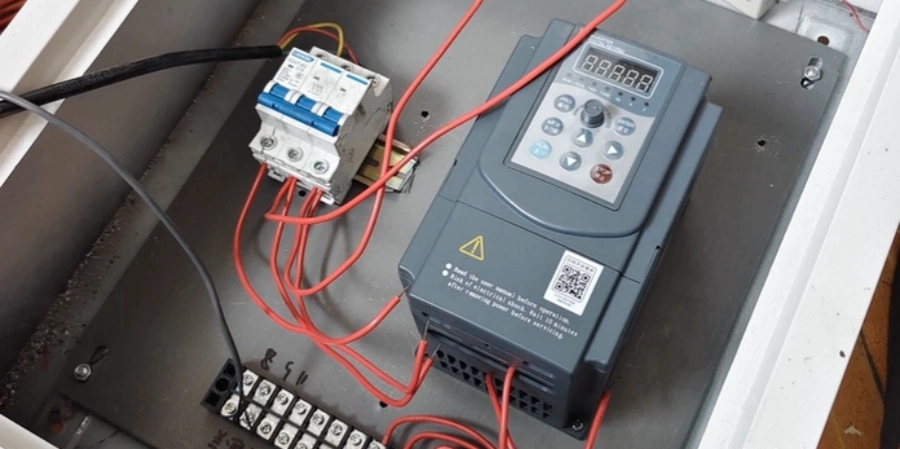 Inside a metal enclosure: gray variable frequency drive, connected with red wires. White three-pole circuit breaker with blue handles above it. Terminal block at bottom.