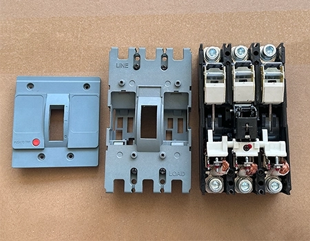 Three MCCB parts laid out on a wooden surface: left - gray front cover with handle and red indicator; middle - bottom half of the cover marked LINE and LOAD (combines with left piece to form the complete cover); right - fully assembled internal mechanism with contacts, arc chutes, and terminals, typical SKD setup ready for final customer assembly.