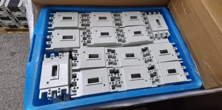 A blue plastic tray holding multiple gray molded case circuit breaker (MCCB) covers arranged in neat rows inside a cardboard box. Each cover has terminal openings, mounting holes, and a small window slot, ready for warehouse storage or shipment.