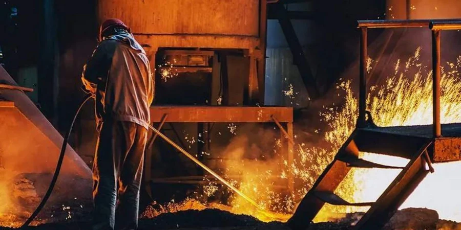 A worker in a red hard hat, silver protective suit, and gloves uses a long pole to manage glowing orange molten metal with sparks flying in an industrial furnace setting.