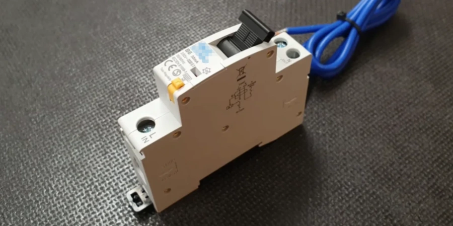 A white single-module residual current breaker with overcurrent protection (RCBO) on a gray surface. Features a yellow test button, black toggle switch in ON position, blue neutral flying lead, and clear markings for N and line connections.
