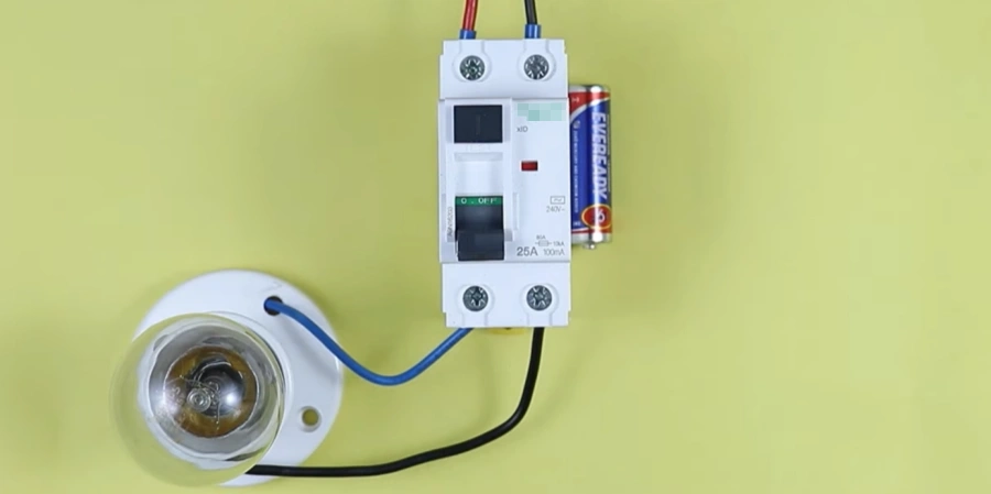 A white residual current circuit breaker (RCCB) connected in a basic test setup on a green background. Red and black wires enter the top, a blue wire connects to a light bulb at the bottom, and an AA battery is wired in to demonstrate leakage current detection and protection.