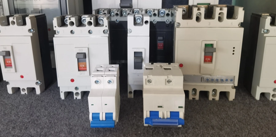 A lineup of low-voltage circuit breakers arranged by size on a gray surface: two small blue-handled miniature circuit breakers (MCBs) in the front, followed by mid-size white MCCBs, and larger beige and black molded case circuit breakers in the back, showing the full progression from residential MCBs to industrial MCCBs.