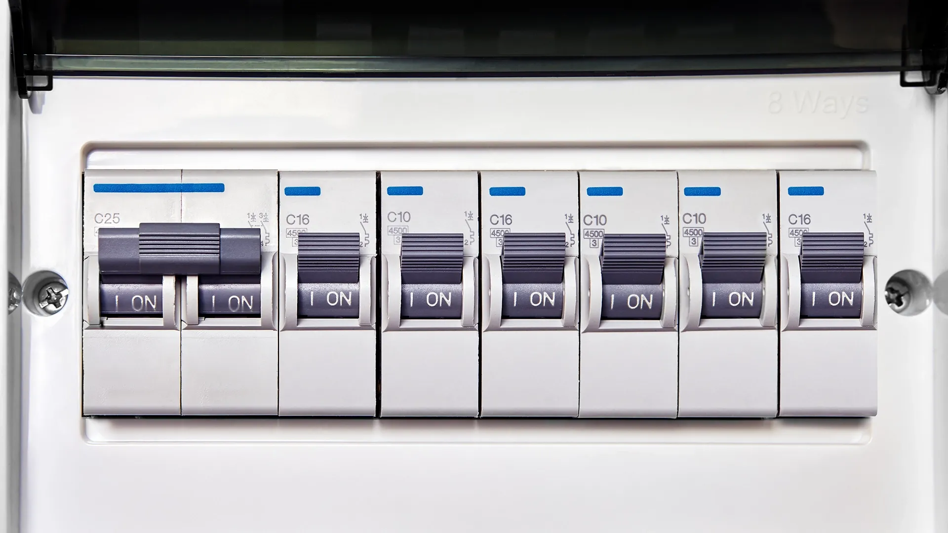 A close-up of an 8-way consumer unit with seven single-pole miniature circuit breakers (MCBs) mounted on a DIN rail. Ratings visible are C25, C16, C10, C16, C10, C10, and C16, all switched to ON position with blue identification flags on top.