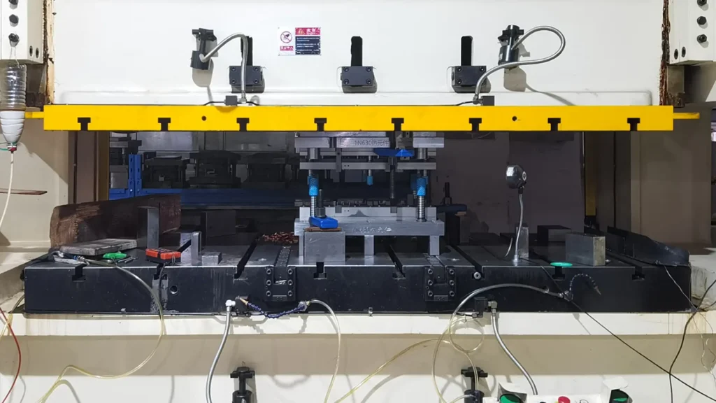 Industrial stamping machine with a yellow safety bar at the top, black base connected to cables and pneumatic tubes, blue tools on the press bed, and control buttons including green start and red stop at the bottom.