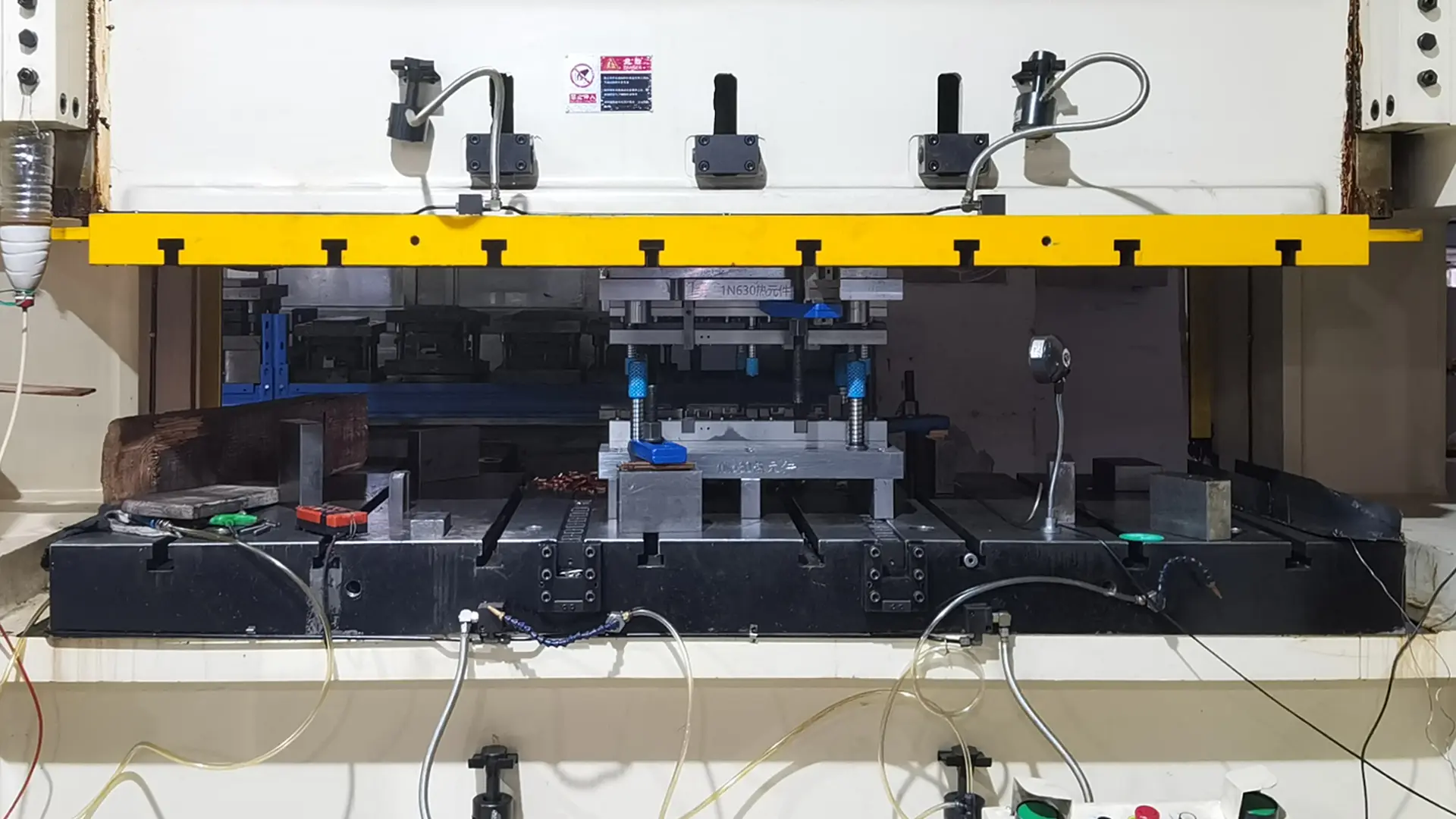 Industrial stamping machine with a yellow safety bar at the top, black base connected to cables and pneumatic tubes, blue tools on the press bed, and control buttons including green start and red stop at the bottom.
