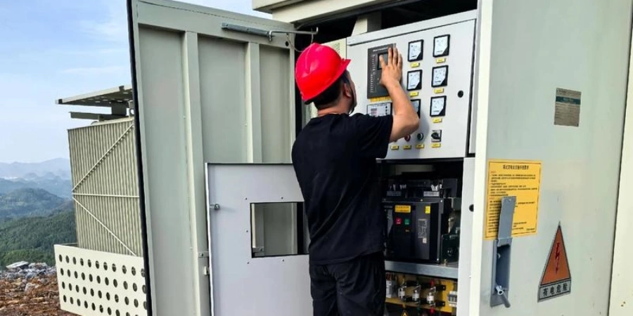 A worker wearing a red hard hat and black shirt adjusts controls on an open electrical panel inside a metal cabinet, located in a high-altitude mountainous region with hills visible in the background.