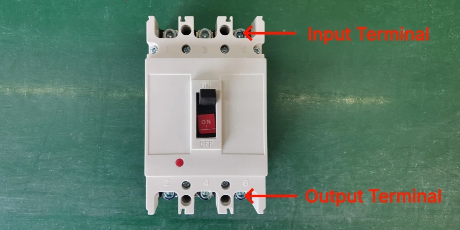 A white three-pole molded case circuit breaker (MCCB) on a green surface, showing the ON/OFF switch in the middle, red indicator light, and numbered terminals 1-6. Red arrows label the top row as Input Terminal and the bottom row as Output Terminal.