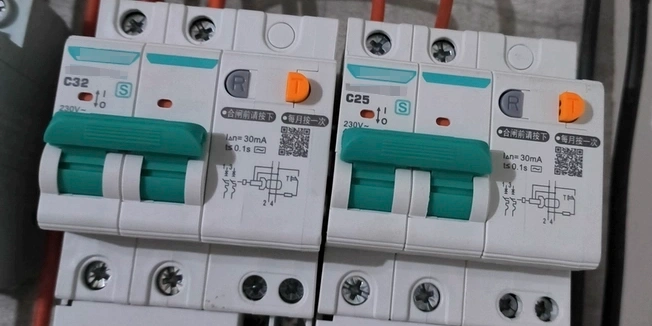 Two white RCBOs mounted side by side. Left one rated C32, right one C25. Both have green handles in ON position, orange test buttons, red indicator lights, and labels showing 230V, 30mA sensitivity.