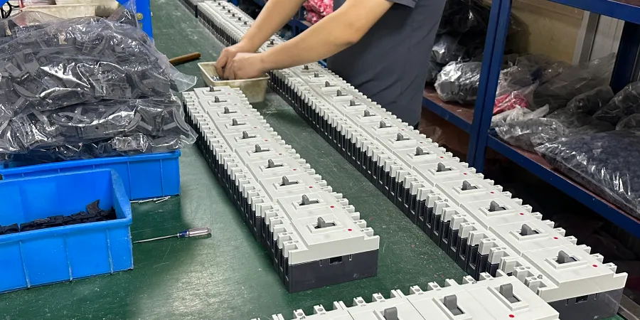A factory worker at a green workbench assembling multiple white and black molded case circuit breakers (MCCBs) lined up in long rows. Tools, parts in blue bins, and packaged components are visible in the background of the manufacturing floor.