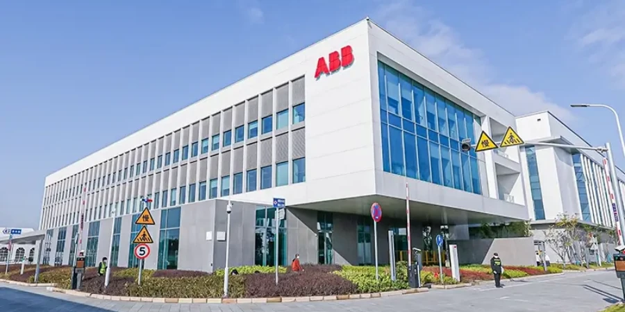 A photograph of a contemporary multi-story industrial building in Xiamen, China, prominently displaying the red ABB logo on its facade. The structure features extensive blue-tinted glass windows, white and gray paneling, and a clean, modern architectural design. In the foreground, a well-maintained landscaped area with shrubs, a paved roadway, traffic signs including a speed limit of 5 km/h and pedestrian warnings, and a few individuals are visible under a clear blue sky. This facility is part of ABB's Xiamen Hub, a key production site for low-voltage switchgear and circuit breakers contributing to the Asia Pacific market.