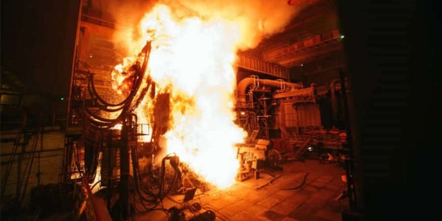 A dramatic photo of a massive fireball and bright arc flash explosion inside an industrial electrical room. Intense orange and white flames burst from equipment, lighting up cables, metal structures, and the floor, showing the dangerous power of an electrical arc blast.