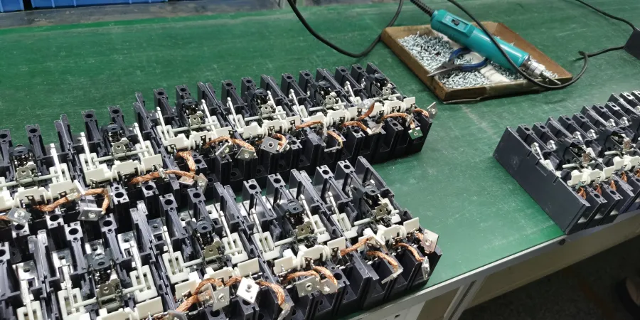 A green industrial workbench displaying multiple partially assembled molded case circuit breakers (MCCBs) arranged in rows, with exposed internal mechanisms including copper braided flexible connections, bimetallic strips, magnetic coils, and contact assemblies. Visible wiring and components indicate an ongoing manual assembly process, accompanied by a tray containing screws and an electric screwdriver, illustrating a production stage focused on constructing the tripping and arc extinguishing mechanisms in low-voltage electrical protection devices.