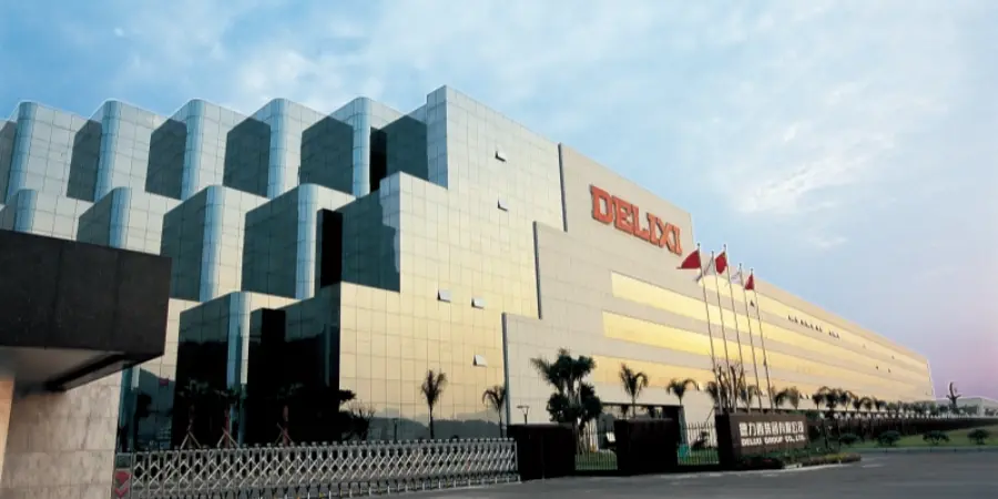 A photograph of the modern Delixi Group headquarters, a multi-story building with a distinctive curved glass facade reflecting the surroundings, prominently featuring the large red 'DELIXI' logo on the upper section. The structure is set against a twilight sky, with palm trees, a gated entrance, flagpoles displaying the Chinese national flag, and well-maintained landscaping in the foreground. Delixi Group, founded in 1984 and originating in Liushi—a leading manufacturer of low-voltage circuit breakers and electrical components, contributing significantly to the Asia Pacific market through its joint venture, Delixi Electric.