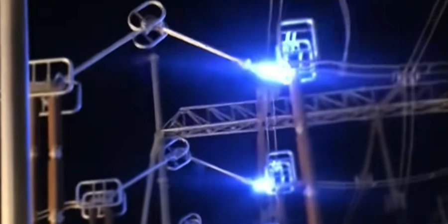 Bright blue-white electrical arcs flash at the contacts of several open disconnect switches mounted on a metal power transmission tower structure at night. The intense light illuminates the insulators and arms against a dark background.