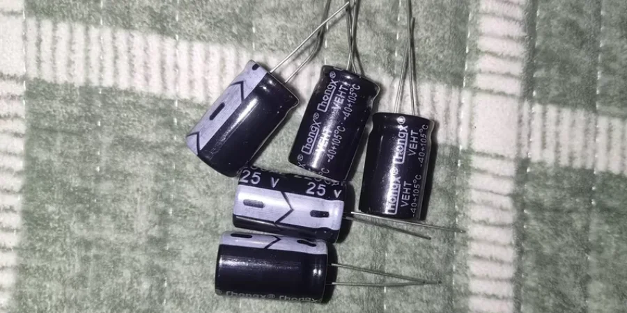 Five black cylindrical electrolytic capacitors with metal leads lying on a green and white checkered fabric background. The labels show ratings like 25V and various capacitance values