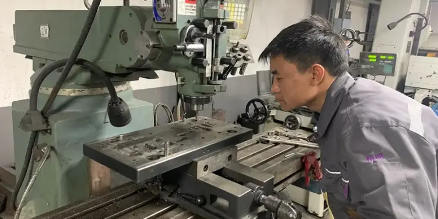 A technician in a gray uniform operates a vertical grinding machine in a manufacturing workshop, closely inspecting the workpiece on the table during precision machining. The setup includes a clamped metal plate, cutting tools, and surrounding equipment, illustrating the in-house mold development process, which is essential for producing high-quality injection molds.