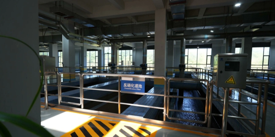 A section of a modern wastewater treatment plant featuring a large open denitrification filter basin with flowing water, enclosed by metal safety railings and marked by a blue sign reading Denitrification Filter. The facility includes tiled basin walls, overhead piping systems, structural support columns, elevated walkways, and large windows allowing natural light, demonstrating an advanced biological process for nitrate removal through anaerobic denitrification in industrial-scale wastewater management.
