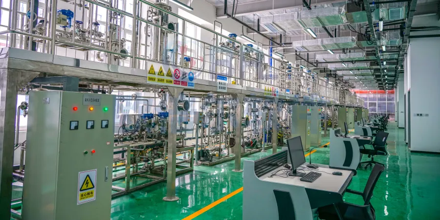 Interior view of a clean petrochemical plant featuring extensive stainless steel piping, valves, reactors, and platforms on multiple levels. Electrical control panels with red and green indicator lights are in the foreground, with operator workstations and computers along the right side on a glossy green floor.