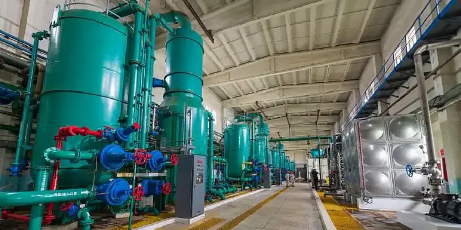 A spacious interior of an oil refinery featuring multiple tall teal-colored pressure vessels and towers, likely adsorption dryers or fractionation columns, interconnected by an extensive network of teal and red piping systems with valves, gauges, and control panels. The facility includes high ceilings, safety-marked walkways, and stainless steel storage tanks, illustrating advanced industrial processes for refining petroleum products in a controlled environment.