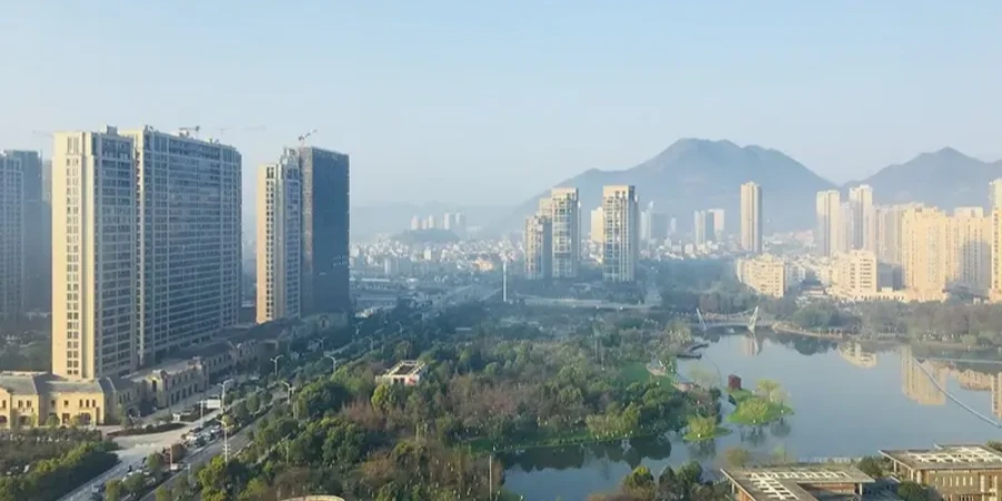 An aerial perspective of Liushi in Yueqing, China, showcasing rapid urban development with clusters of high-rise residential buildings, some under construction with cranes visible, set against a backdrop of hazy mountains. In the foreground, a serene lake reflects the surroundings, bordered by lush green parks, trees, and pathways, illustrating the blend of modern infrastructure and natural scenery in this region renowned as the 'Capital of Electrical Appliances' for its prominence in low-voltage circuit breaker manufacturing.