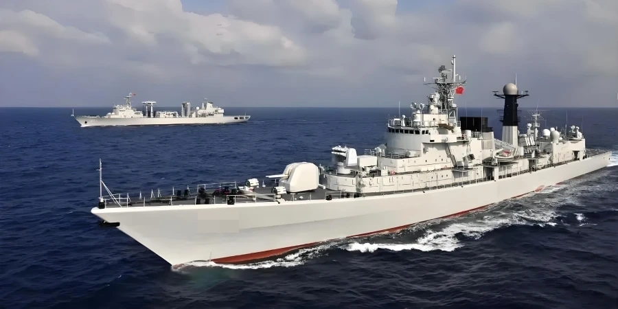 A large white marine ship in the foreground cuts through blue ocean waves under a cloudy sky, with a red flag flying on its mast. Another gray marine ship sails parallel in the background during a marine exercise or patrol.